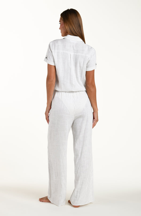 Full body back image of model wearing a pant jumpsuit in a button up collared front with wide pockets, short sleeves, pant pockets, and clean back. Featuring a cream colored linen print. 