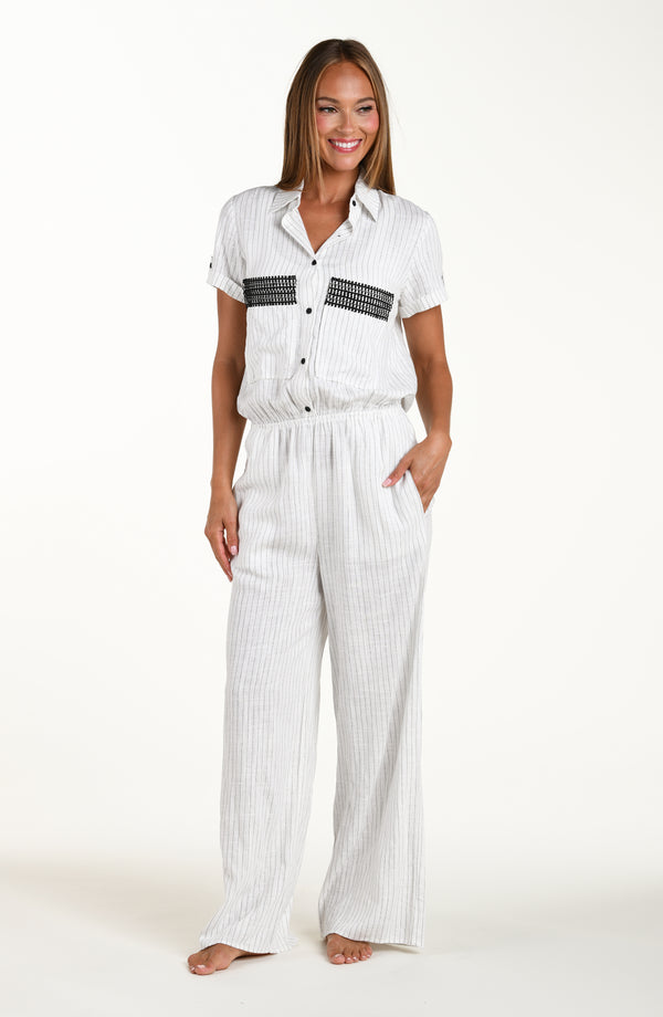 Full body front image of model wearing a pant jumpsuit in a button up collared front with wide pockets adorned with crochet trim, short sleeves, and pant pockets. Featuring a cream colored linen print. 
