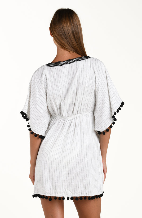 Back image of model wearing a short pull over v-neck caftan with three quarter sleeves adorned with tassel trim. Featuring a cream colored linen print.