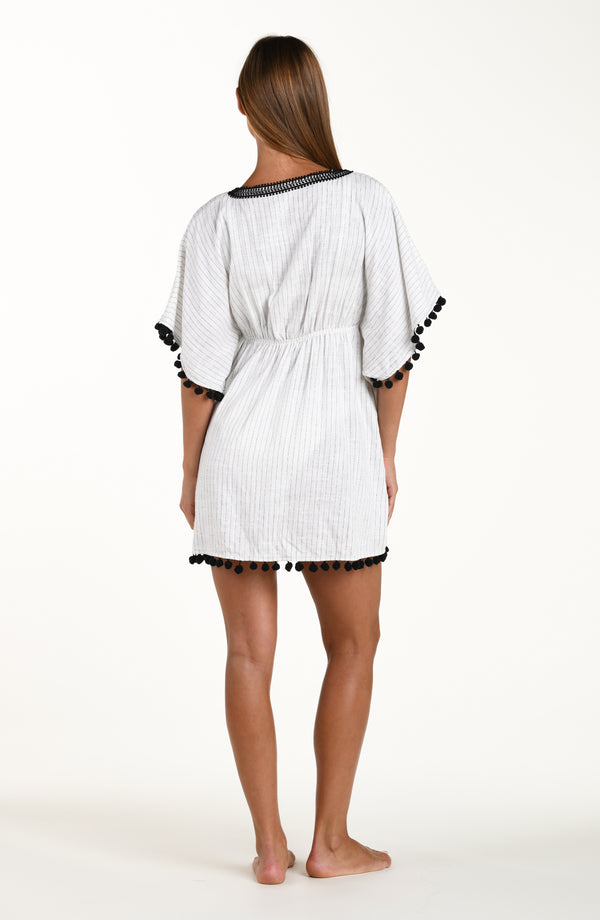 Full body back image of model wearing a short pull over v-neck caftan with three quarter sleeves adorned with tassel trim. Featuring a cream colored linen print.