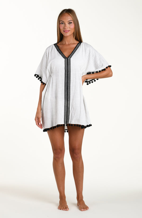 Full body front image of model wearing a short pull over v-neck caftan with three quarter sleeves adorned with tassel trim. Featuring a cream colored linen print.