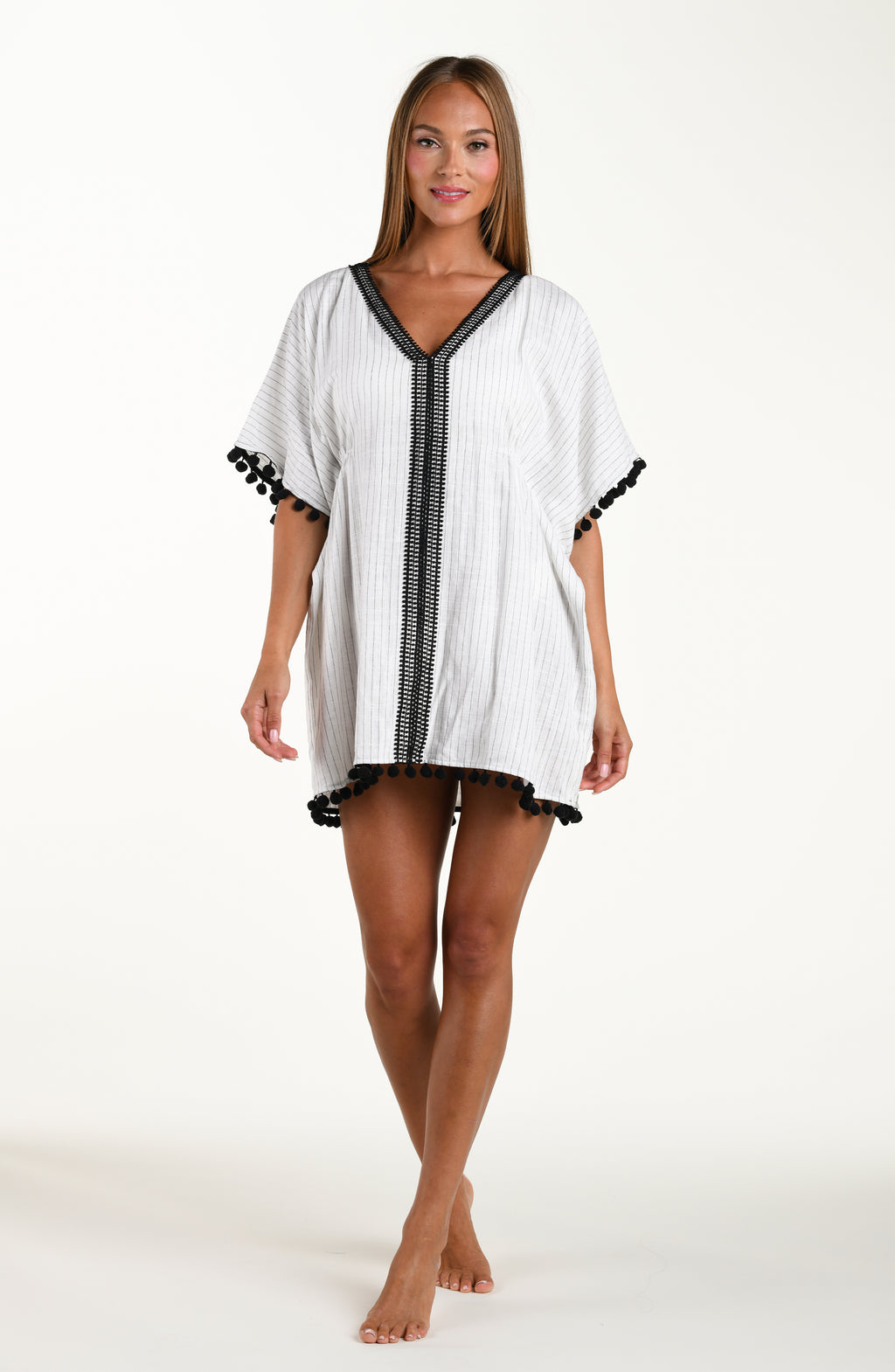 Full body front image of model wearing a short pull over v-neck caftan with three quarter sleeves adorned with tassel trim. Featuring a cream colored linen print.