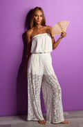 Woman in a white lace romper holding a fan against a purple wall