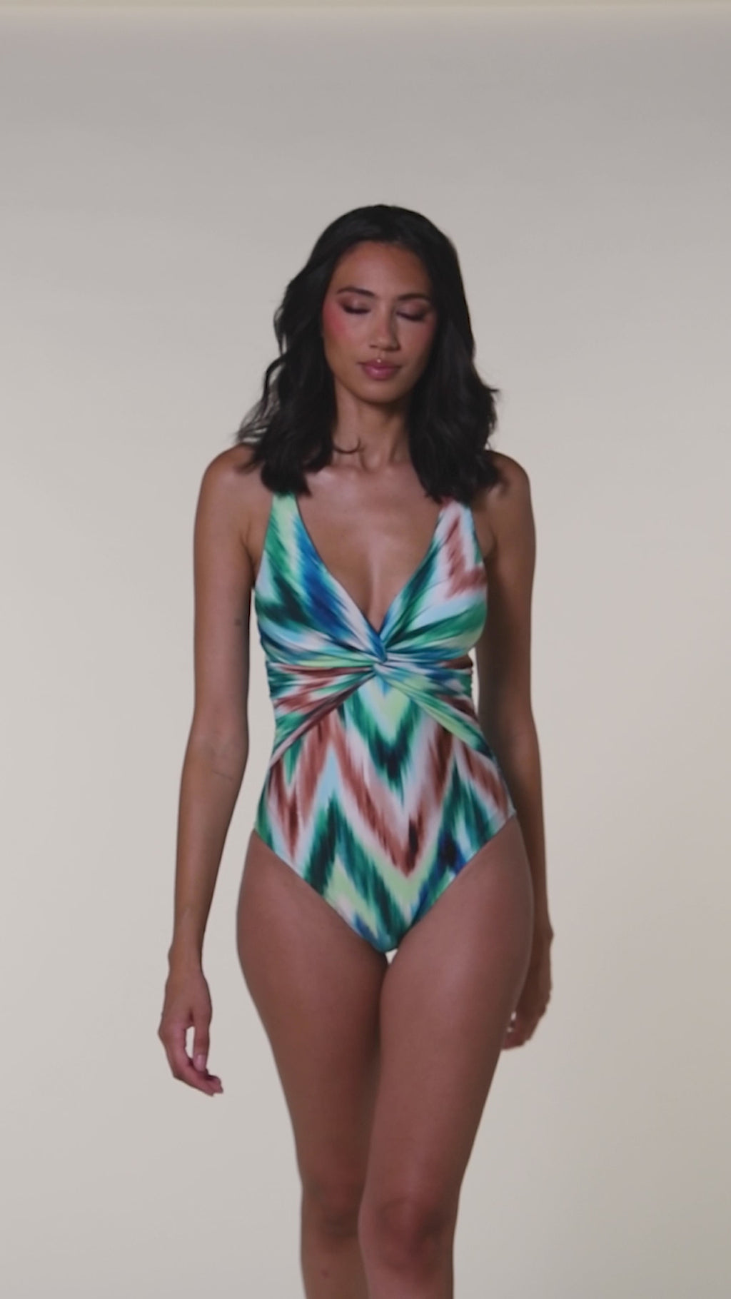 Video of a woman wearing a green multicolored one piece swimsuit against a white background. 