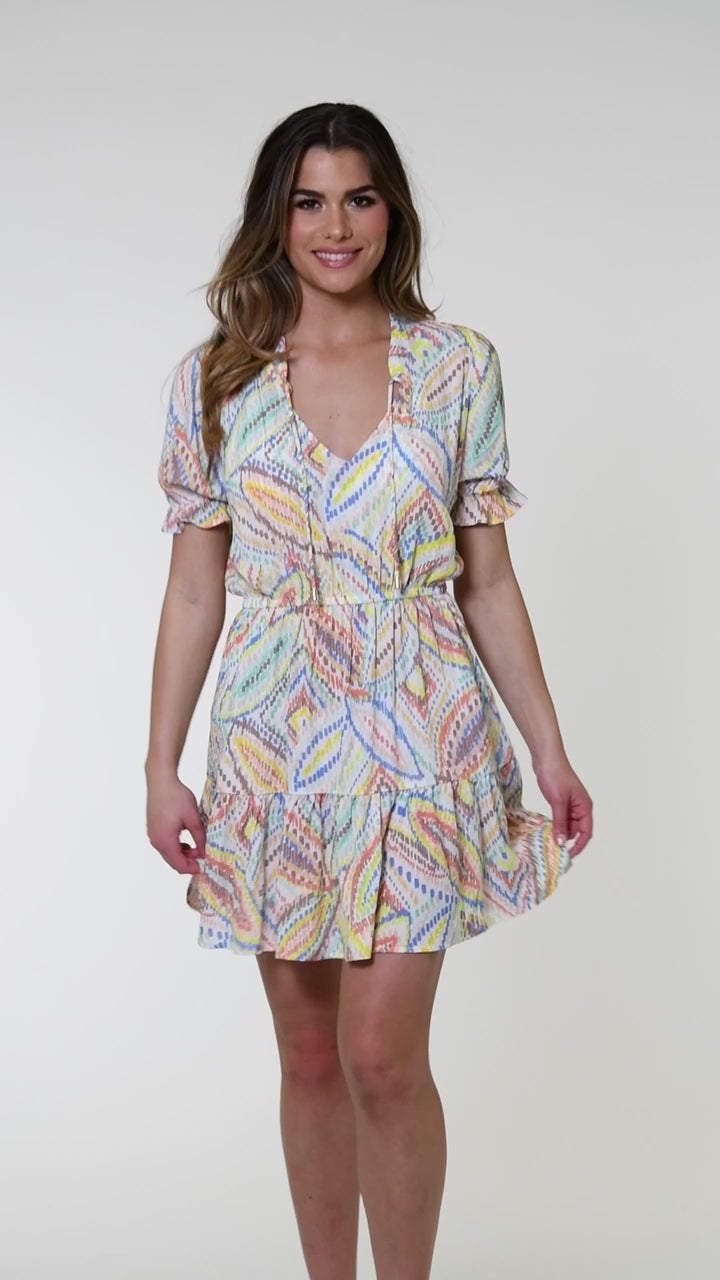 Model is wearing a geometric patterned cover-up dress in pastel shades of pink, blue, and yellow, accented with sporadic glimmers of gold sequin-like details on a white background.