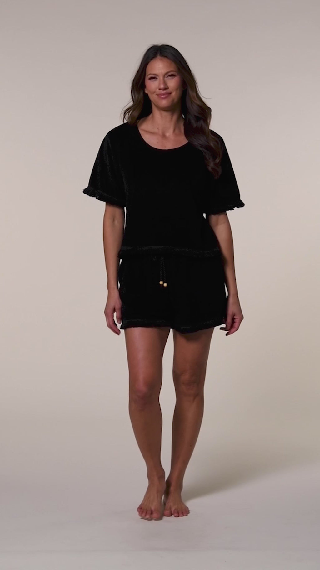 Video of a woman wearing a black short sleeve top and back shorts with drawstring and gold buttons against a white background