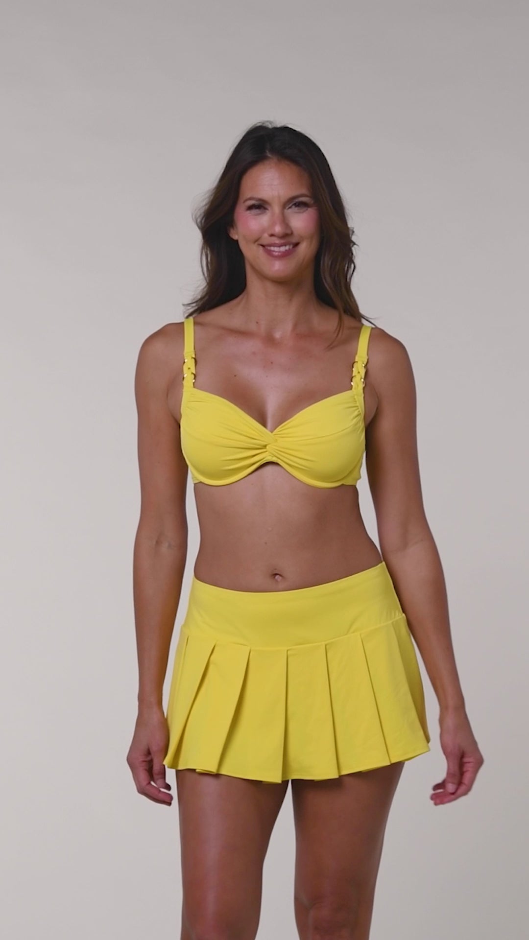 Video of a woman wearing a yellow bikini top and matching swim skirt against a white background. 
