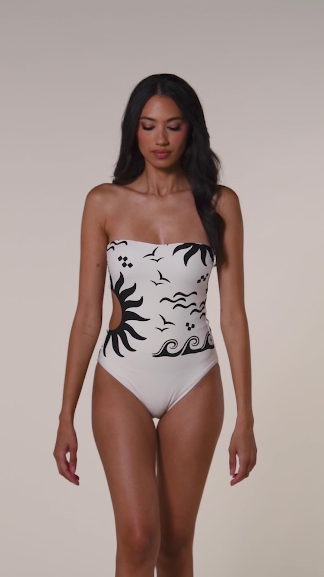 Video of a woman wearing a white one piece swimsuit with black designs on a white background. 
