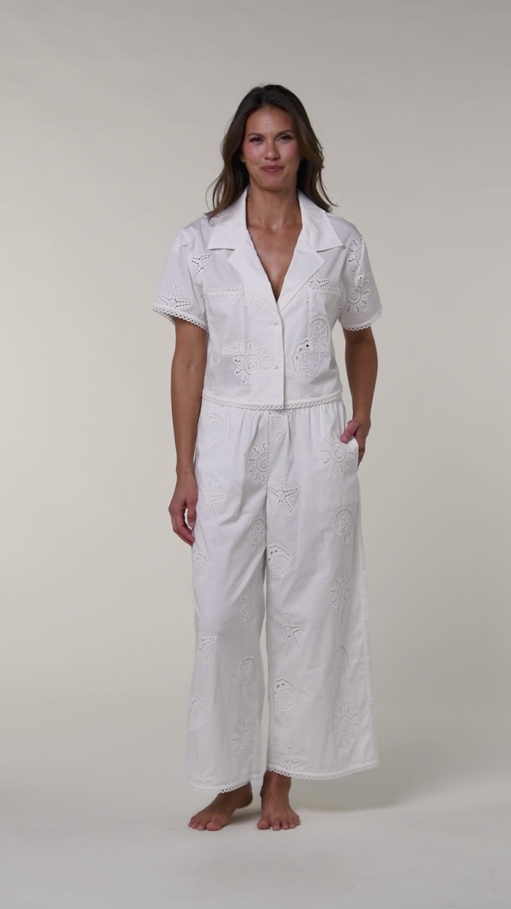 Video of a woman wearing white cover up pants and a white bikini top against a white background. 
