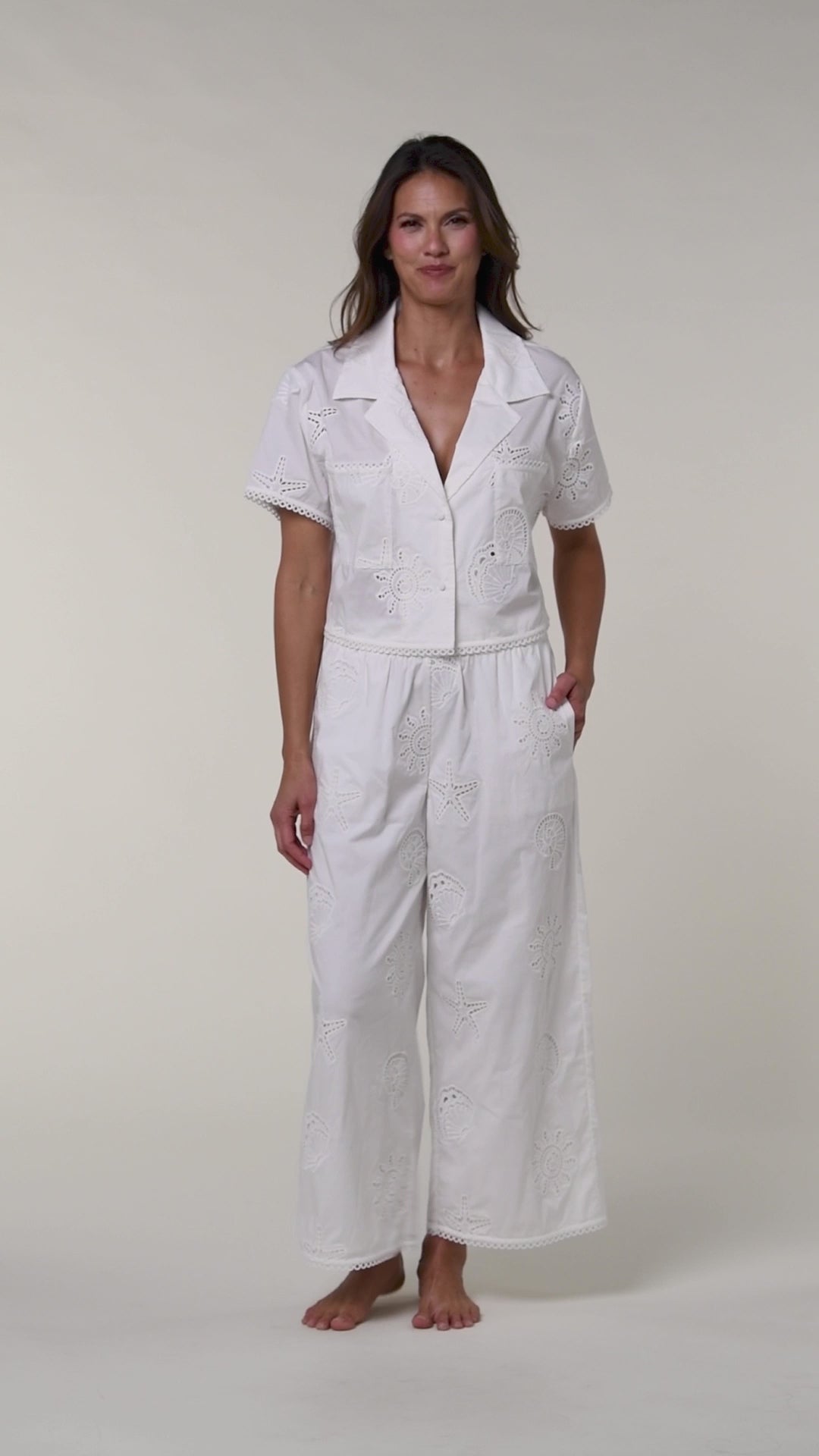 Video of a woman wearing white cover up pants and a white bikini top against a white background. 