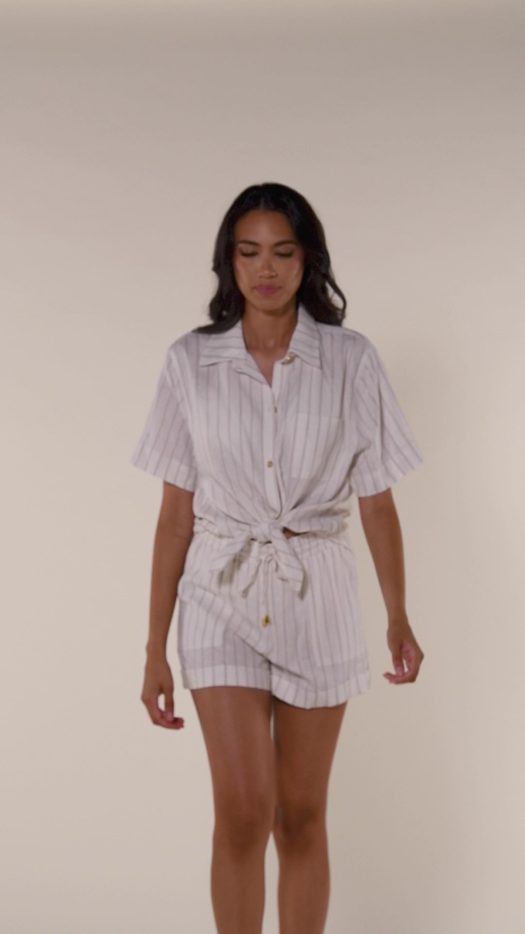 Video of model wearing a cream striped colored short-sleeve cover-up shirt with button front, chest pocket, and tie-front waist. It's paired with matching beach shorts.