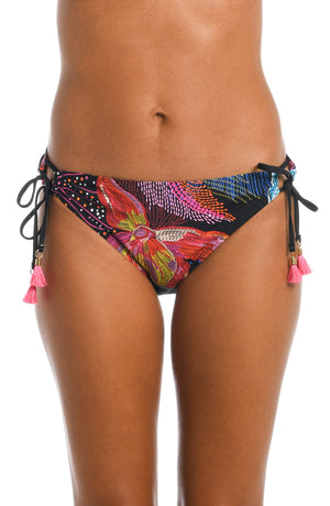 
            
                Load image into Gallery viewer, Model is wearing a shiny multicolored tropical printed side tie hipster bottom from our Sunlit Soriee collection!
            
        