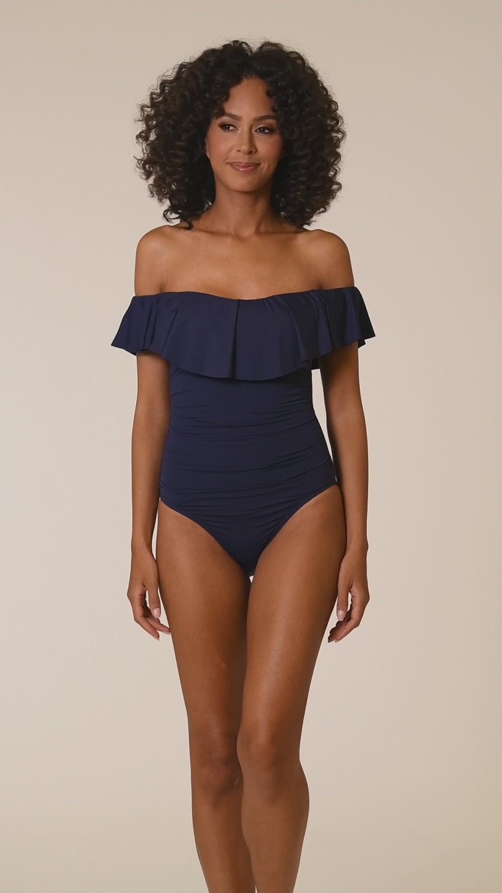 Video of model wearing an indigo (navy blue) colored off-the-shoulder one-piece swimsuit featuring a flounce ruffle neckline, ruched bodice, and moderate rear coverage.