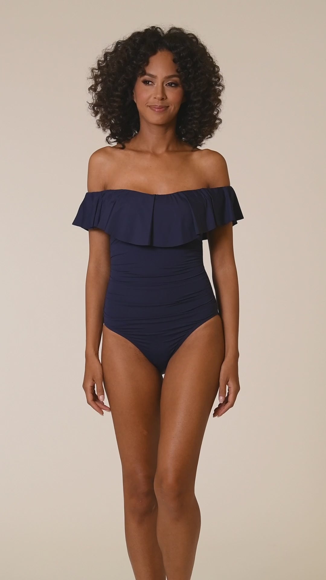 Video of model wearing an indigo (navy blue) colored off-the-shoulder one-piece swimsuit featuring a flounce ruffle neckline, ruched bodice, and moderate rear coverage.
